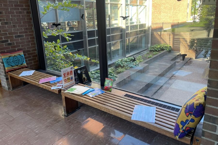 Photograph of a bench on the library bridge with pillows and reading materials. An outside view is visible through the window behind the bench. 