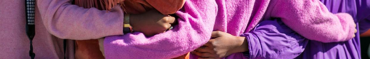 Photo of four people's backs with their arms around each other. Two people are wearing pink coats, one is wearing an orange coat and the last, purple.