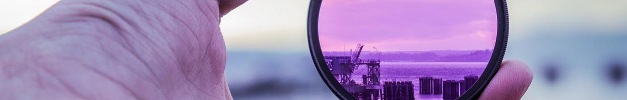 A white person's hand holding a circular pink filter, through which a distant landscape is visible.